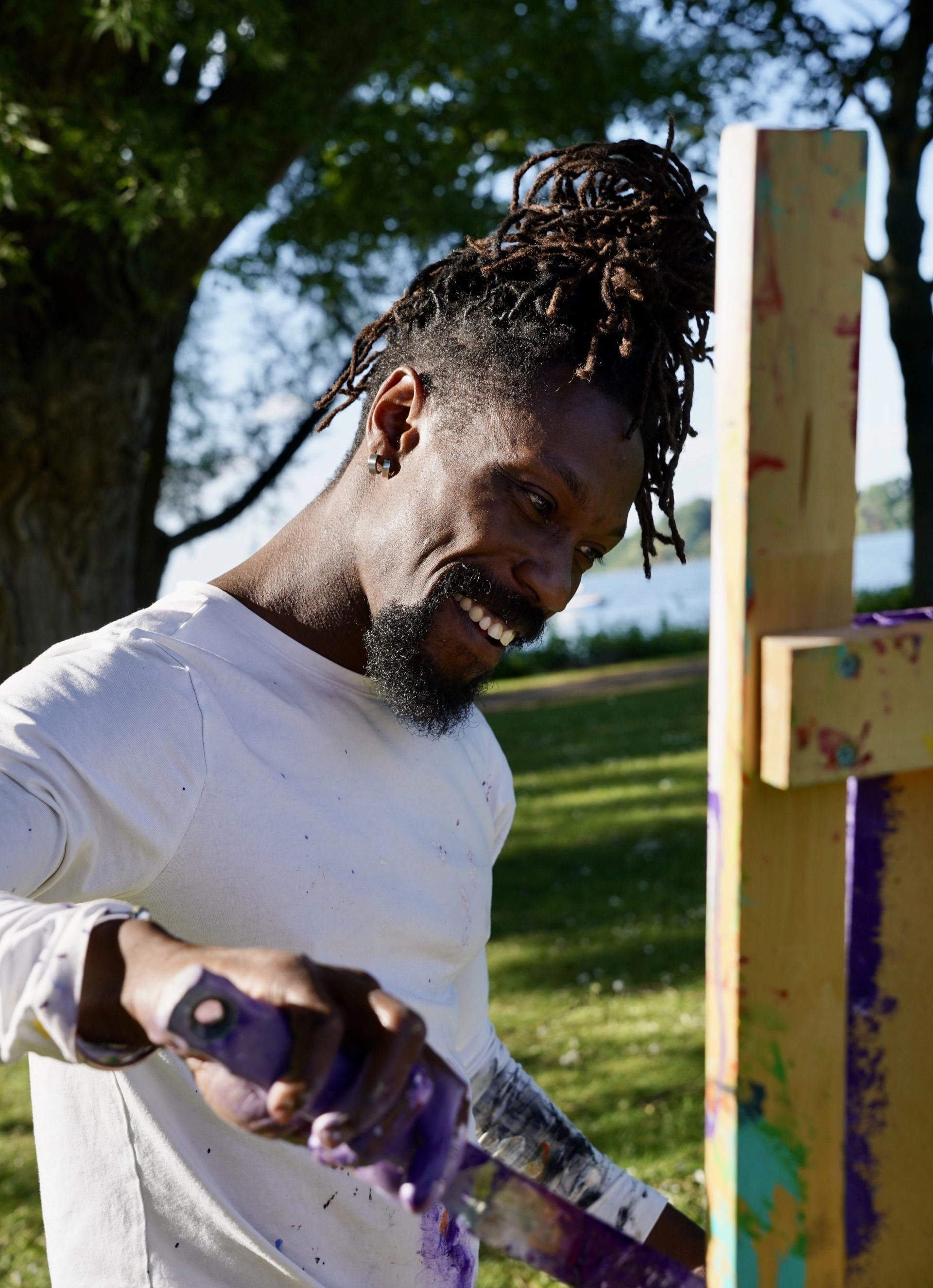 A smiling man with dreadlocks paints a colorful canvas outdoors on a sunny day, surrounded by trees and grass.